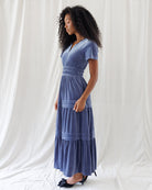 Side profile showing the defined high-waist smocking, classic short-sleeve cut, and sweeping tiered A-line silhouette of the soft blue velvet dress.