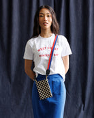 Woman wearing a white t-shirt with text, blue pants, and a checkered bag against a dark curtain background
