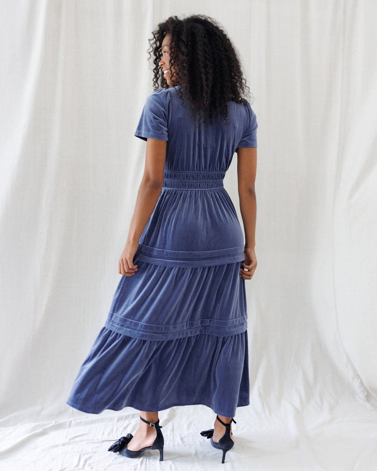 Back view highlighting the gathered bodice, flexible smocked waistband, subtle puff shoulder seams, and flowing multi-tiered velvet maxi skirt.