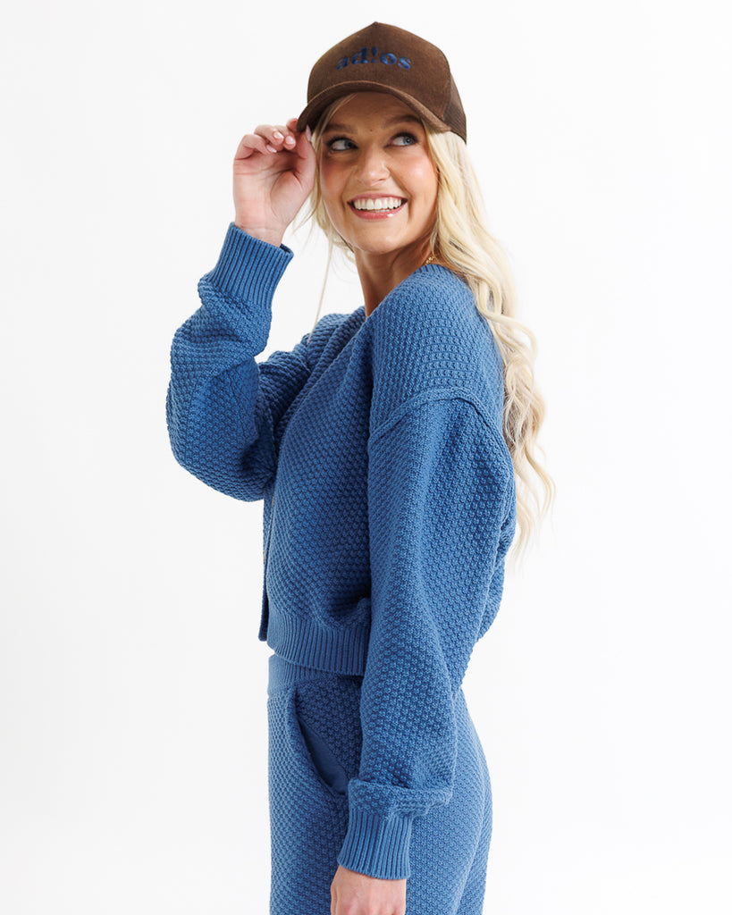 Woman wearing a blue textured sweater and pants set with a brown cap on a white background