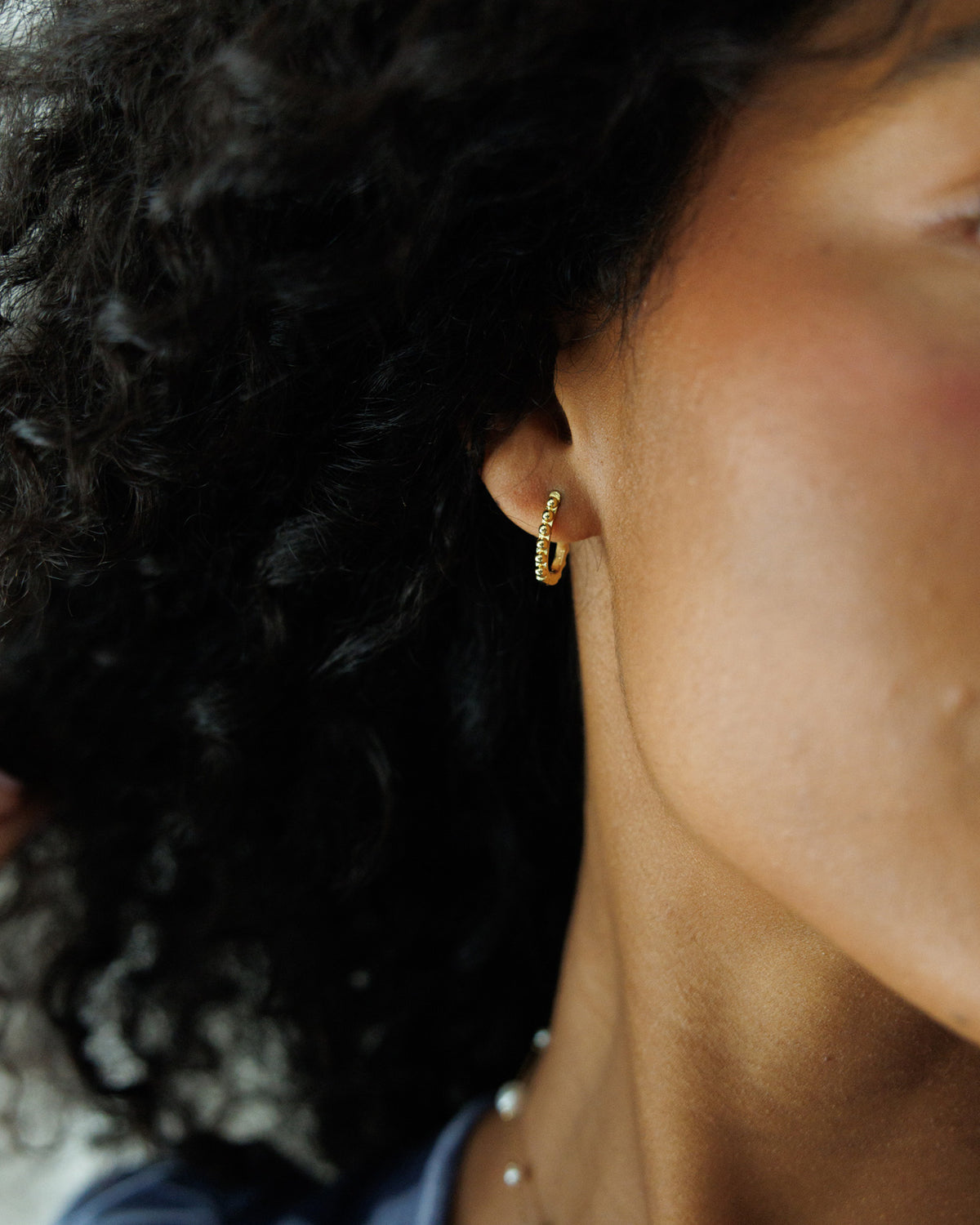 A close-up side profile of a person with curly dark hair wearing a small gold hoop earring with tiny clear stones, along with a pearl necklace.