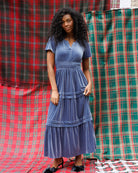 A woman wearing a dusty blue tiered midi dress with short sleeves and a smocked waist, standing in front of a red and green tartan plaid backdrop.