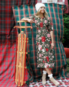 A woman wearing a green floral midi dress, white knit beanie, white socks, and beige ankle boots stands beside a wooden sled in front of red and green plaid fabric backdrops.