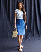 Woman wearing a white floral top and blue skirt holding a yellow bag against a dark background