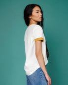 Woman wearing a white t-shirt with yellow trim and blue jeans against a teal background
