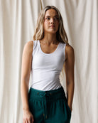 A woman with long blonde hair wearing a white scoop-neck tank top and teal joggers, shown from the waist up against a plain white studio background.