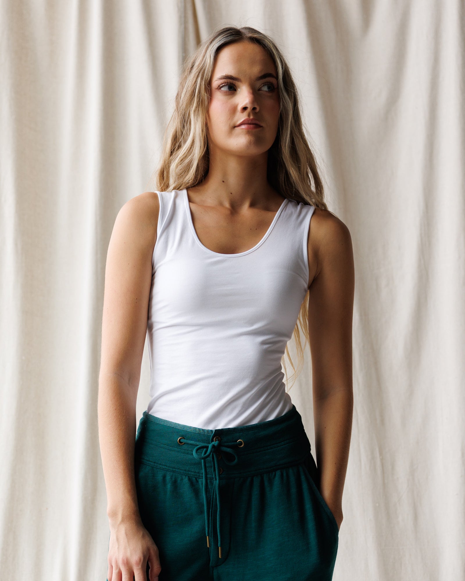A woman with long blonde hair wearing a white scoop-neck tank top and teal joggers, shown from the waist up against a plain white studio background.