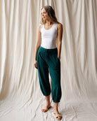 A woman with long blonde hair wearing a white scoop-neck tank top and teal joggers, against a fabric background.