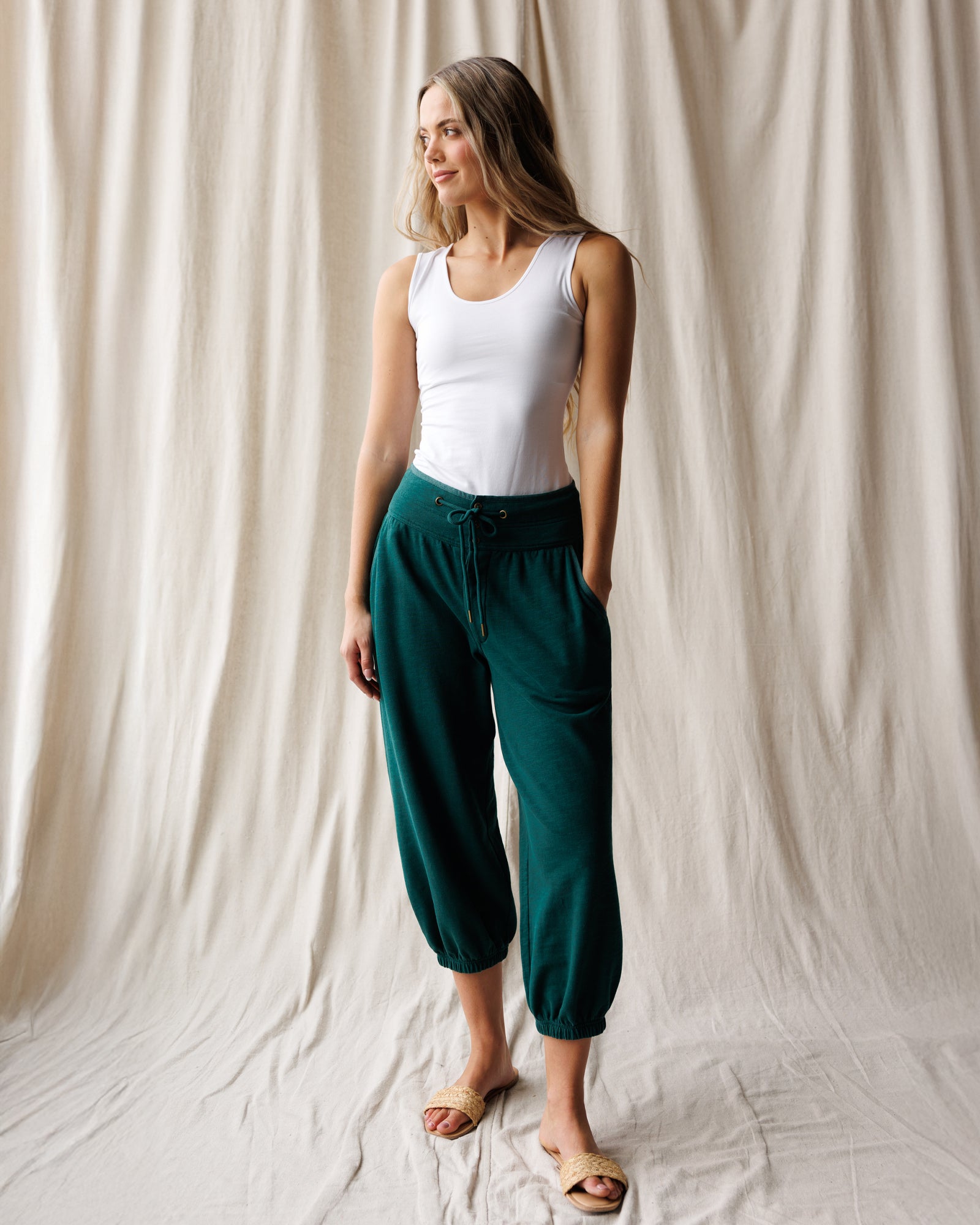 A woman with long blonde hair wearing a white scoop-neck tank top and teal joggers, against a fabric background.