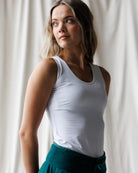 A woman with long blonde hair wearing a white scoop-neck tank top and teal joggers, shown from the waist up against a plain white studio background.