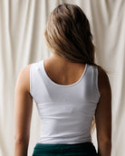 A woman with long blonde hair wearing a white scoop-neck tank top and teal joggers, shown from the waist up against a plain white studio background.