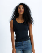 A woman with curly black hair wearing a black scoop-neck tank top and blue jeans, standing against a plain white studio background.