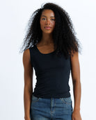A woman with curly black hair wearing a black sleeveless scoop-neck tank top and blue jeans, standing against a plain white studio background.