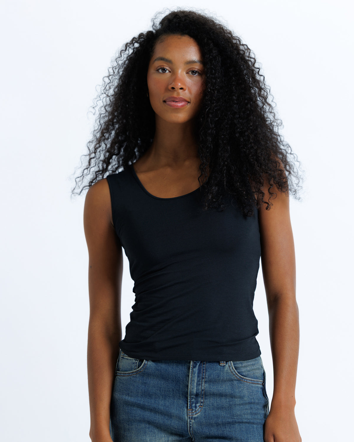 A woman with curly black hair wearing a black sleeveless scoop-neck tank top and blue jeans, standing against a plain white studio background.