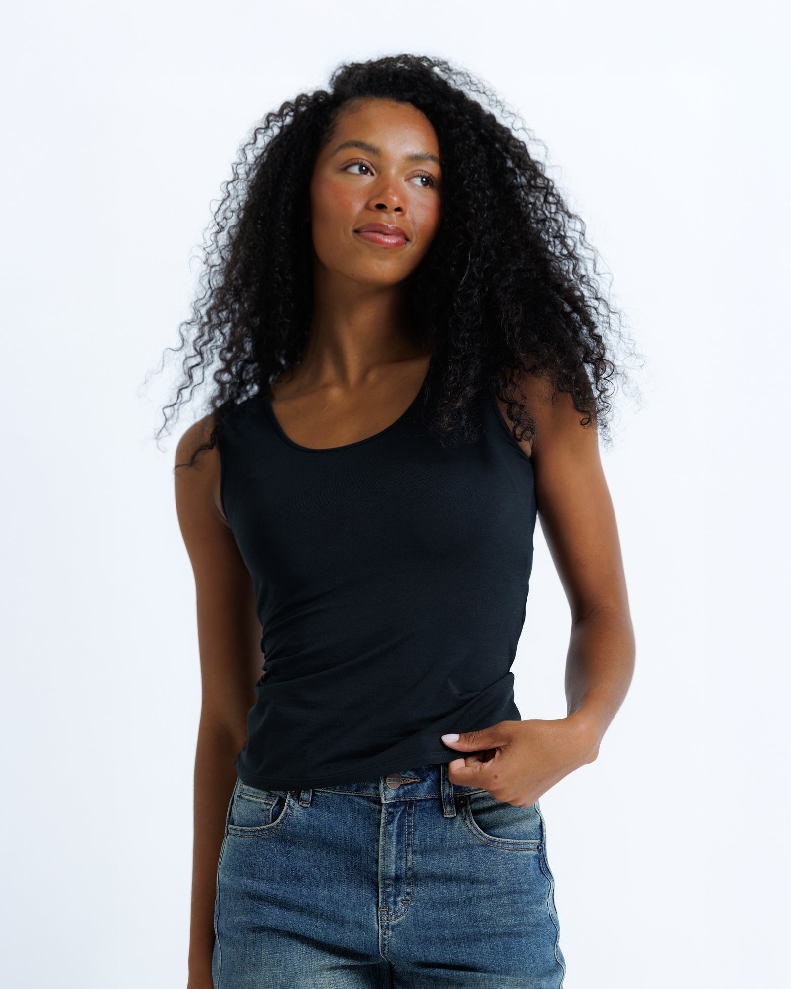 A woman with curly black hair wearing a black scoop-neck tank top and blue jeans, standing against a plain white studio background.