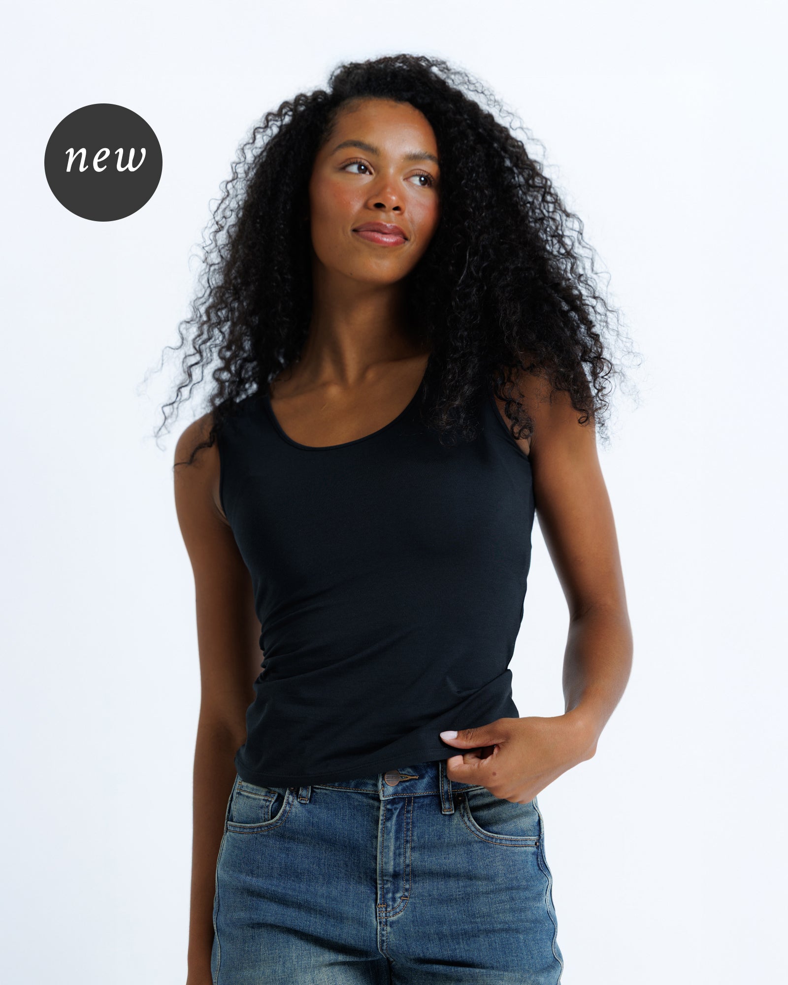 A woman with curly black hair wearing a black scoop-neck tank top and blue jeans, standing against a plain white studio background.