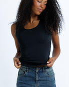A woman with curly black hair wearing a black scoop-neck tank top and blue jeans, shown from the waist up against a plain white studio background.