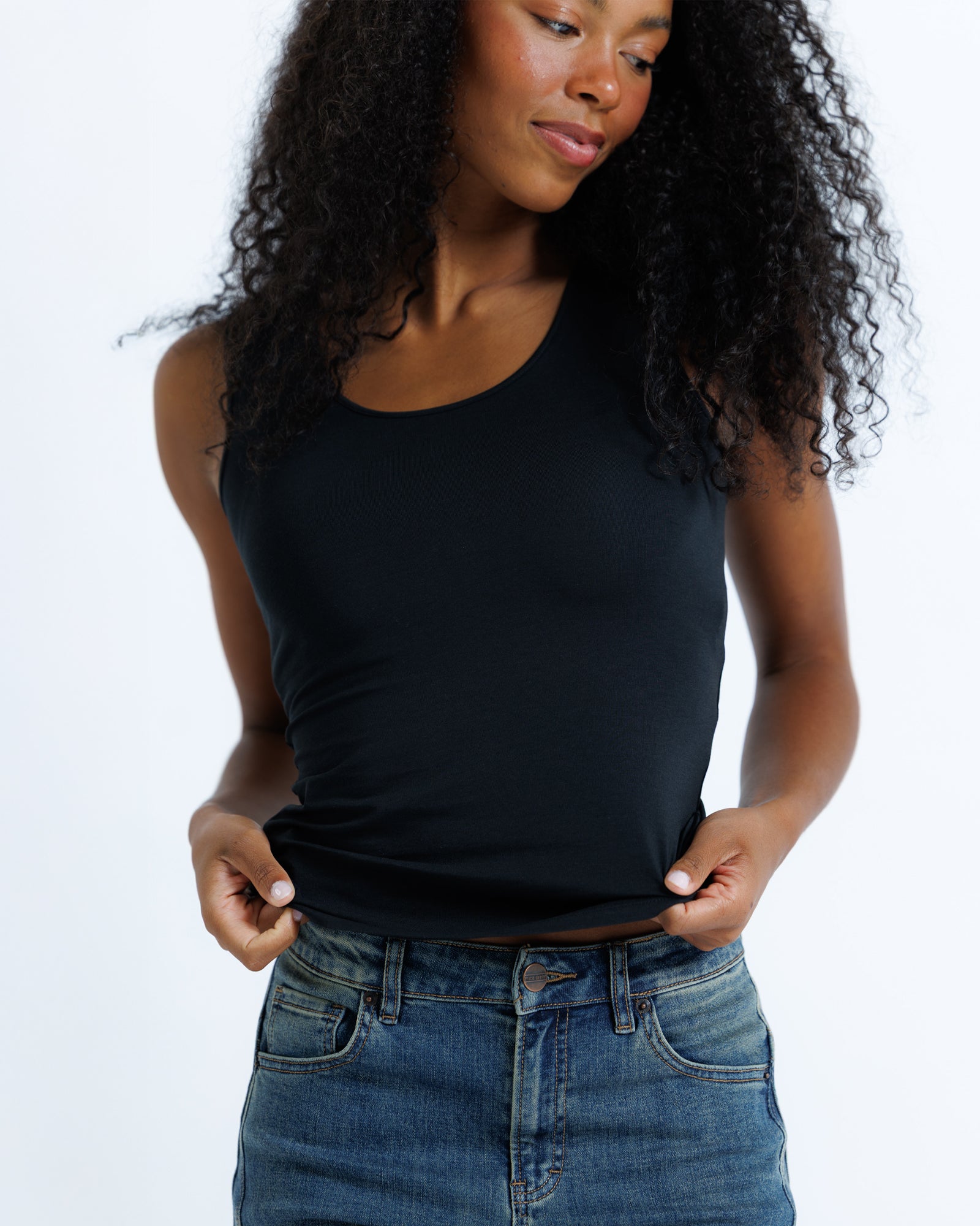 A woman with curly black hair wearing a black scoop-neck tank top and blue jeans, shown from the waist up against a plain white studio background.