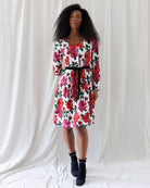A woman modeling a white long-sleeve knee-length dress with a vibrant red and pink floral print and a black waist tie, paired with black ankle boots, set against a draped white fabric backdrop.