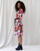 A woman stands in profile wearing a knee-length white dress with a bold red, pink, and green floral print and a black waist tie, paired with dark ankle boots, against a draped white fabric backdrop.