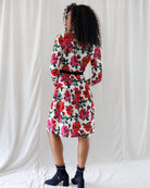 A model is shown from the back wearing a white knee-length dress with a vibrant red and pink floral print, a black waist belt, and black ankle boots, standing against a white draped fabric backdrop.