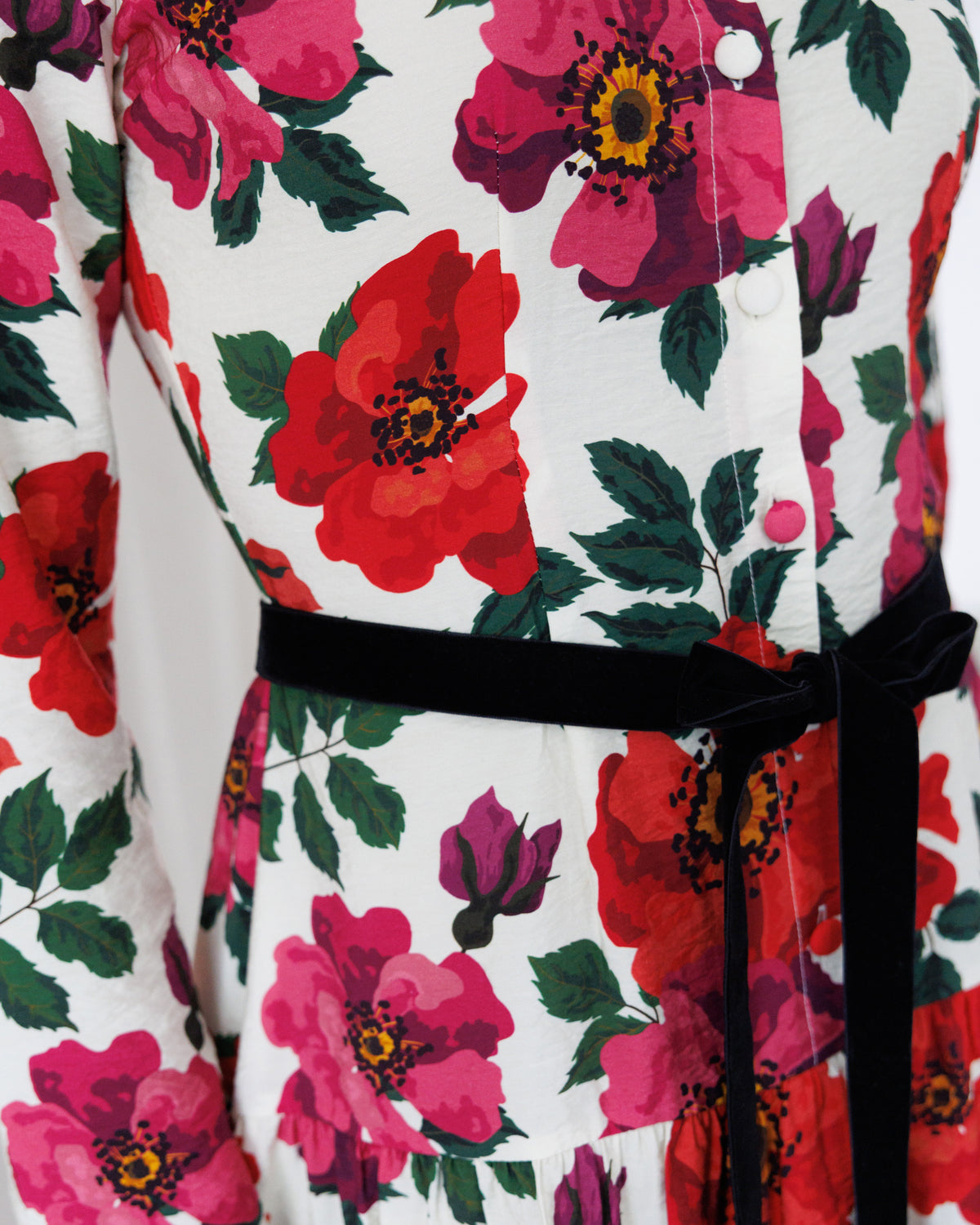 A close-up of a white button-front garment with a bold floral print featuring large red and pink flowers with green leaves, accented by a black velvet tie belt at the waist.