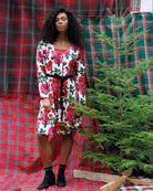 A woman stands in front of red and green tartan plaid fabric backdrops, wearing a white knee-length dress with a bold red and pink floral print, green leaves, and a black waist tie. She is wearing black ankle socks and black boots, standing next to a small artificial Christmas tree.