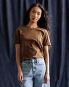 A woman wearing a brown short-sleeve crewneck t-shirt and light blue jeans, standing against a dark blue textured fabric backdrop.