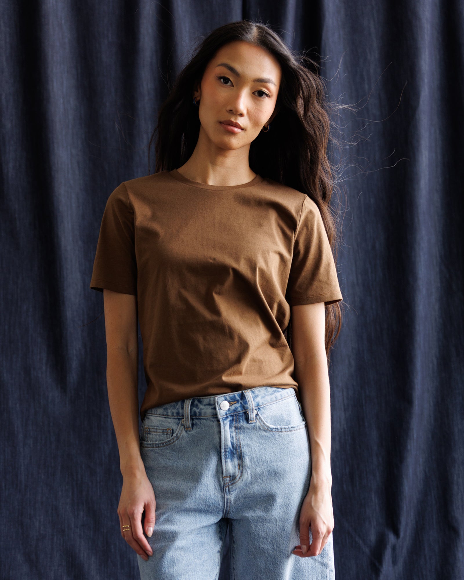 A woman wearing a brown short-sleeve crewneck t-shirt and light blue jeans, standing against a dark blue textured fabric backdrop.