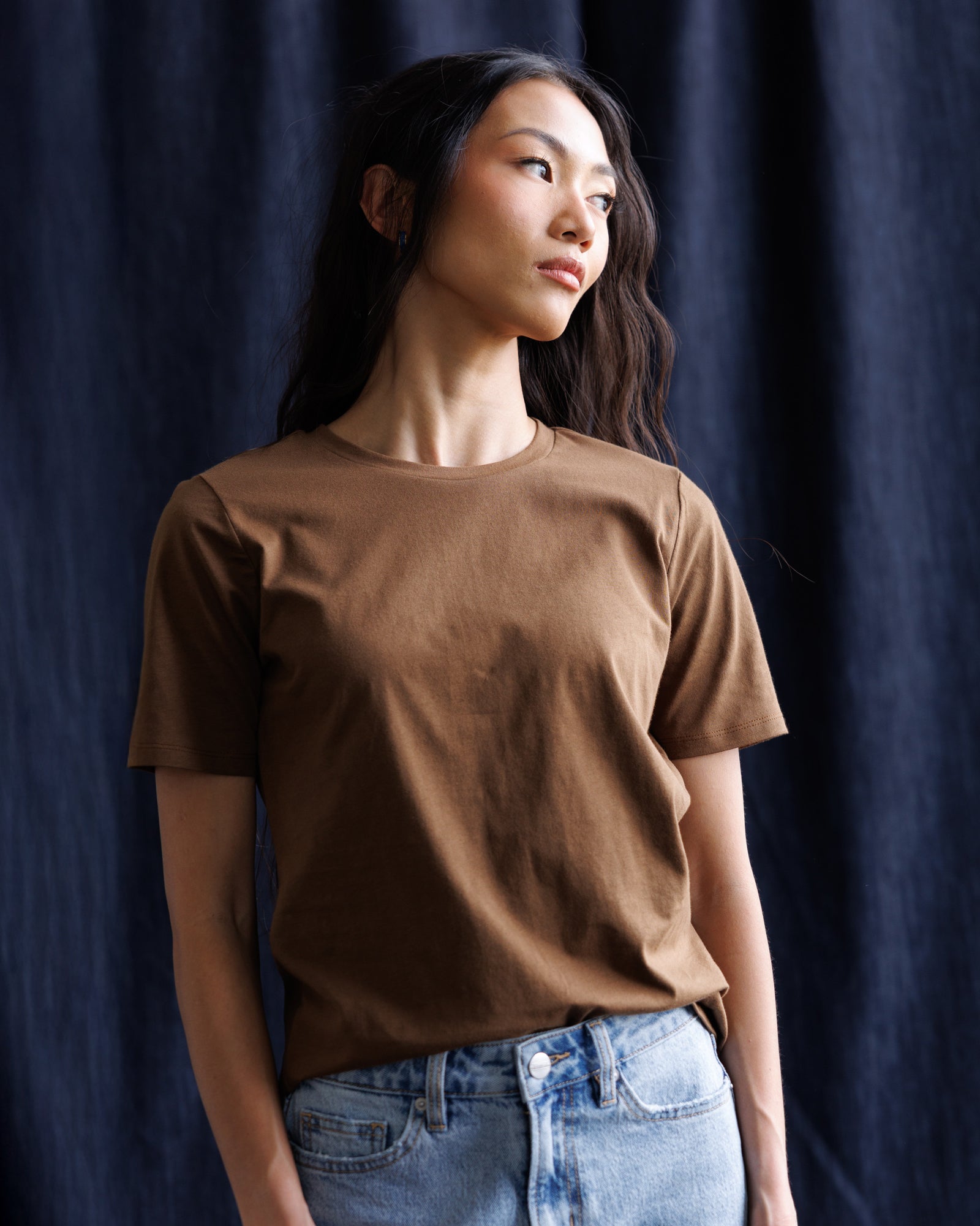 A woman wearing a brown short-sleeve crewneck t-shirt and light blue jeans, standing against a dark navy blue curtain backdrop.