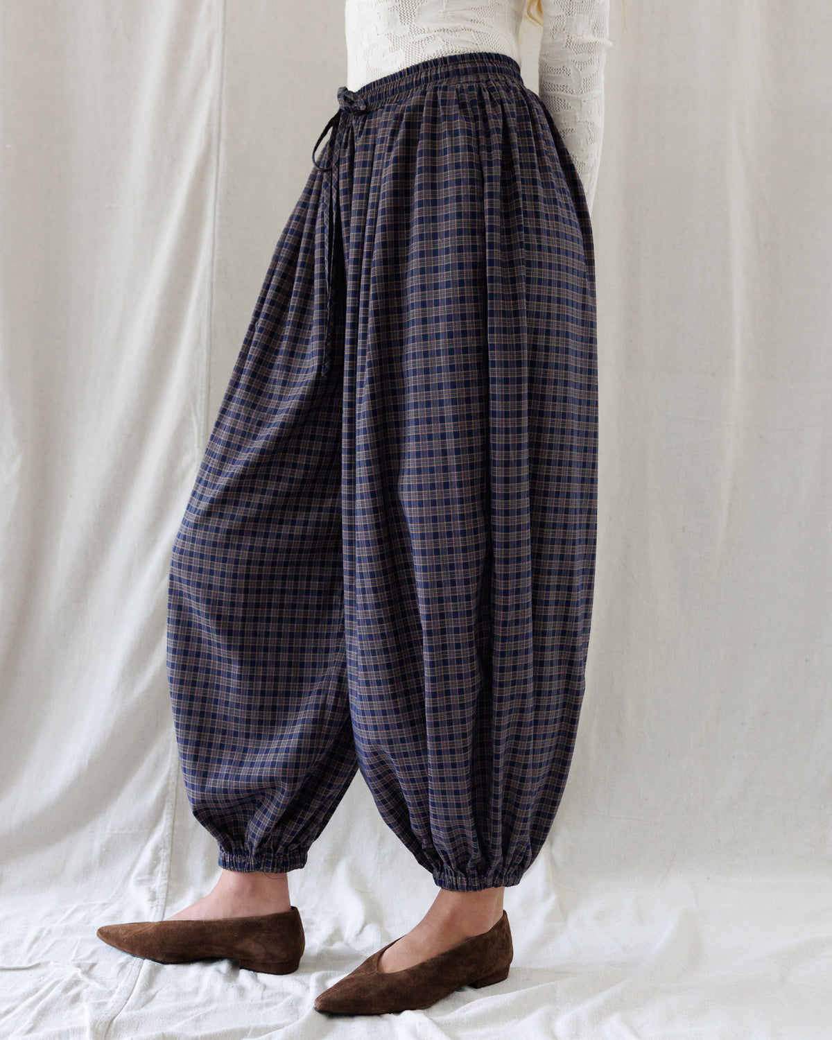 A person wearing loose, balloon-style harem pants with a dark navy and brown plaid pattern, paired with brown flat shoes, standing against a white fabric backdrop.
