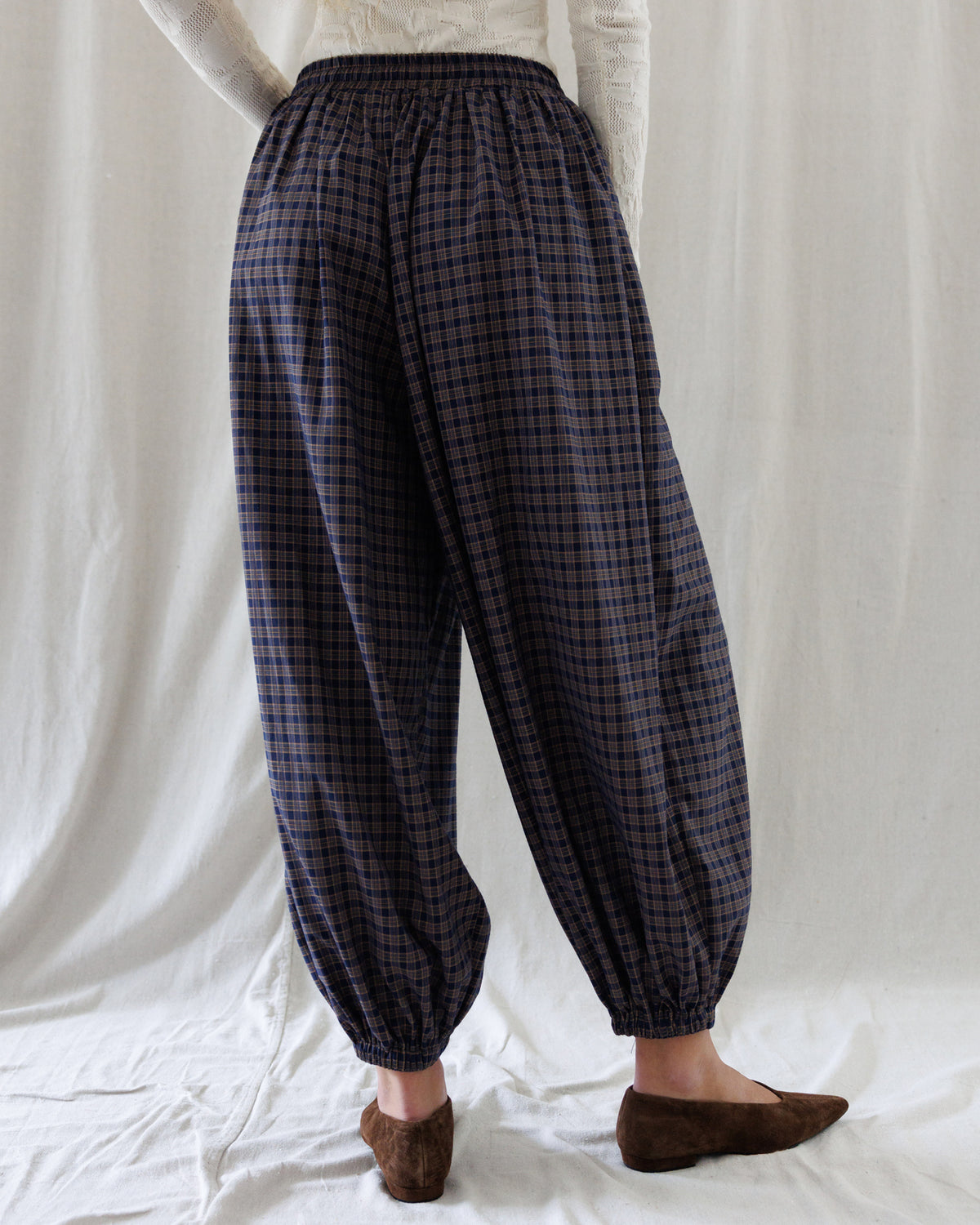 A person shown from the back wearing loose, balloon-style pants with a dark navy and brown plaid pattern, paired with a white top and brown slip-on shoes, standing against a white fabric backdrop.