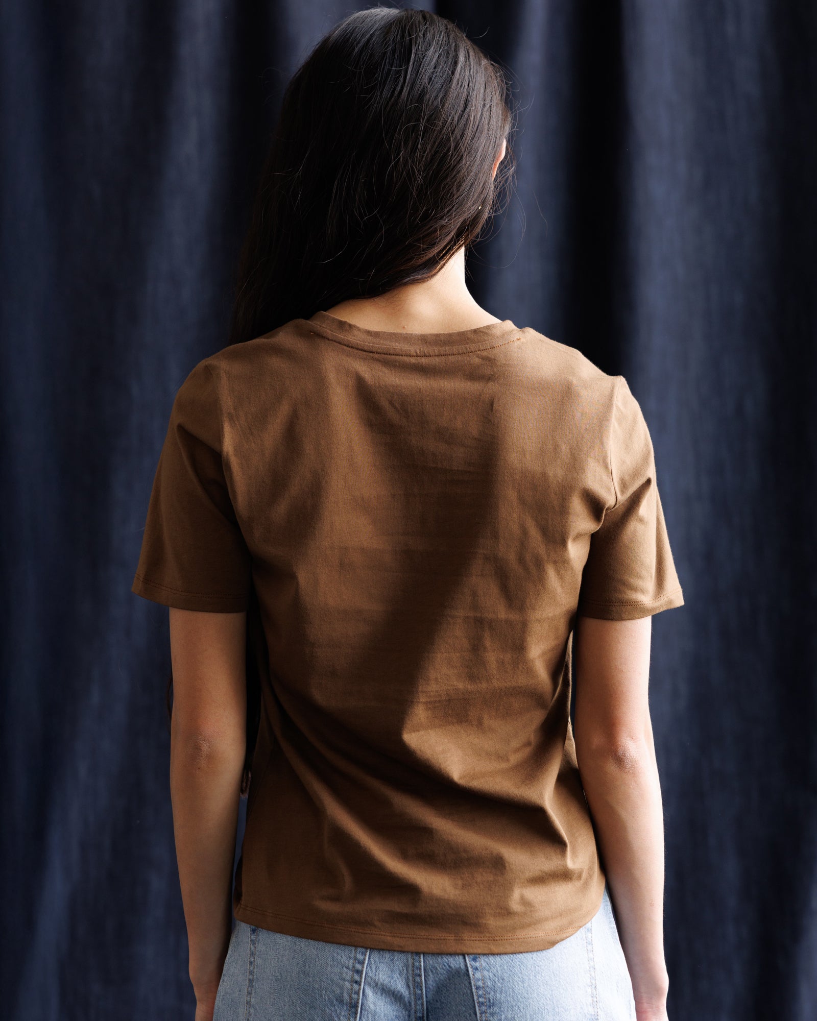 A person viewed from behind wearing a brown short-sleeve crew-neck t-shirt and light blue jeans, standing against a dark navy blue textured fabric backdrop.