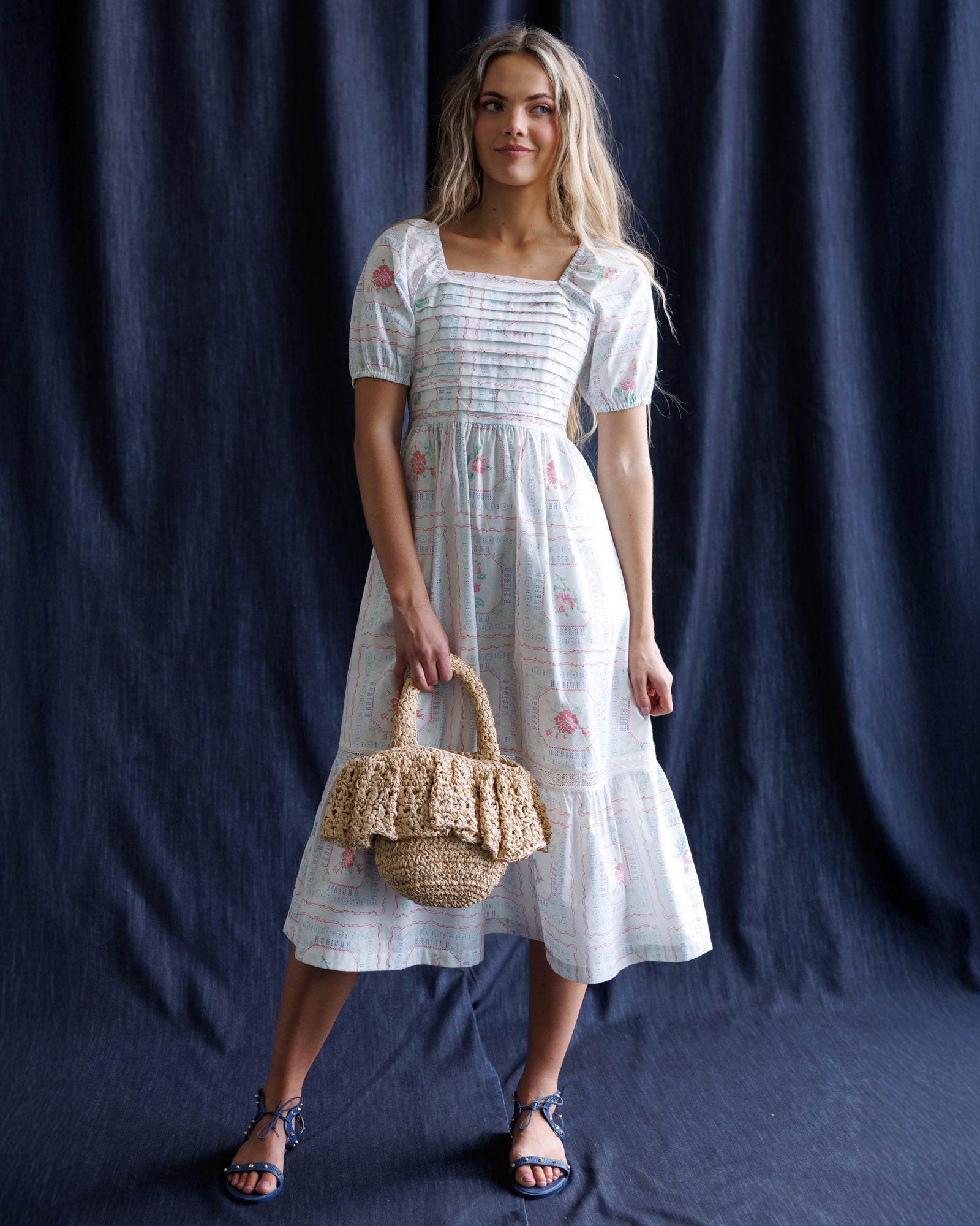 Woman in a white dress holding a woven bag against a dark blue curtain