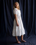 Woman wearing a white dress with floral patterns against a dark blue curtain.