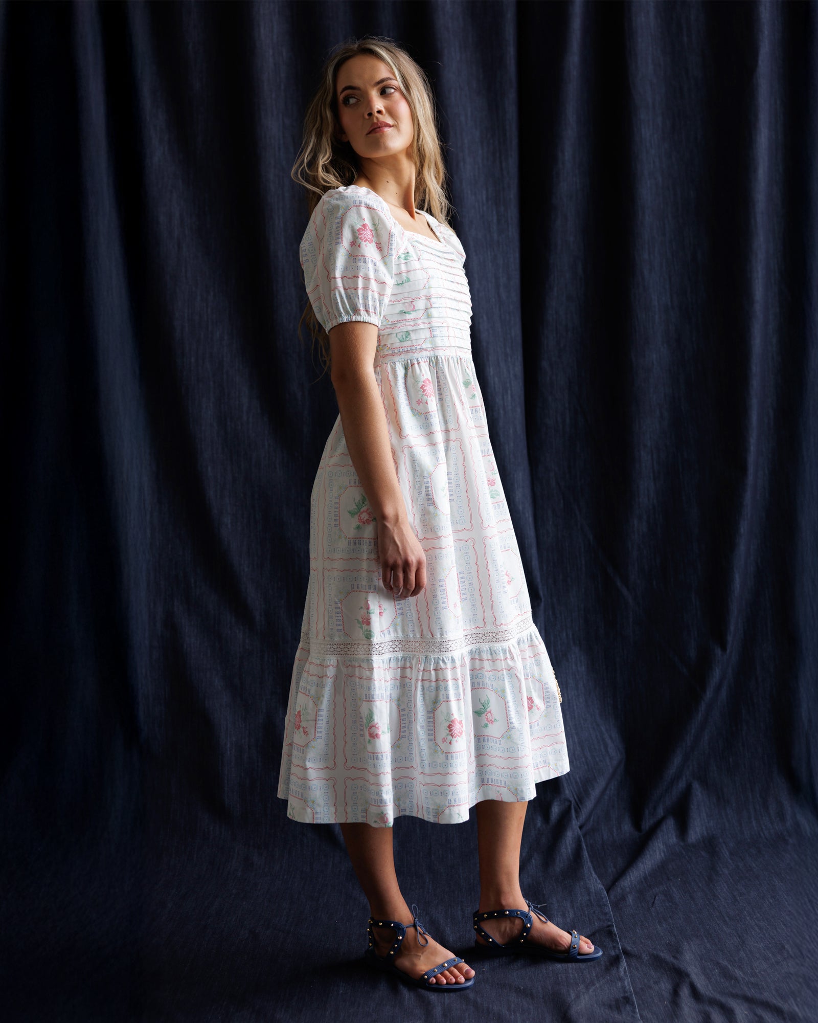 Woman wearing a white dress with floral patterns against a dark blue curtain.