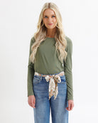 Woman wearing an olive green long-sleeve top and blue jeans with a patterned belt on a white background