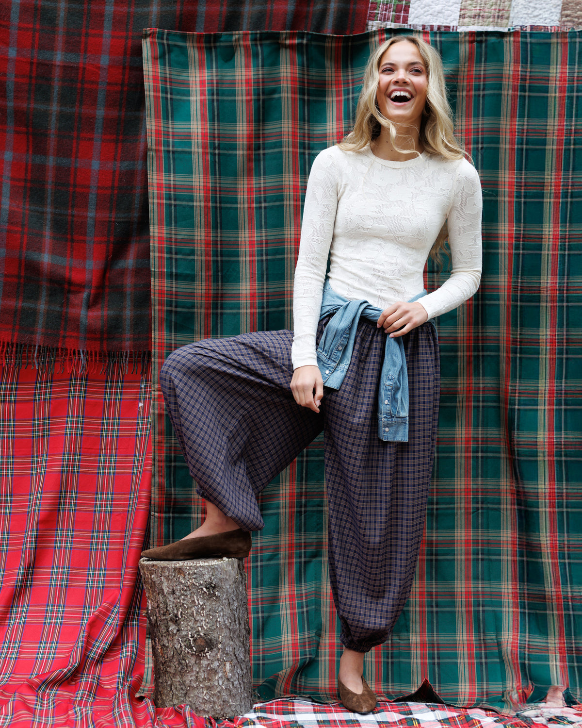 A woman stands in front of plaid fabric backdrops, wearing a cream ribbed long-sleeve top, dark gray checkered wide-leg pants, and a light blue denim shirt tied around her waist.