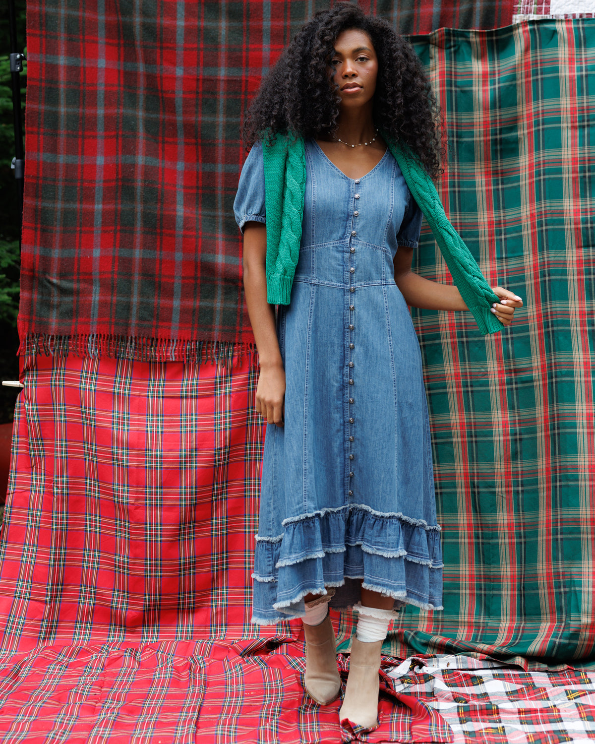 A woman stands in front of red and green plaid fabric backdrops, wearing a blue denim midi dress with short puff sleeves, a V-neck, front buttons, and a tiered hem. She has a bright green scarf draped over her shoulders and is wearing beige ankle boots.