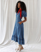 A woman modeling a blue denim midi dress with short puff sleeves, side pockets, and a red neck scarf, standing against a draped white fabric backdrop.