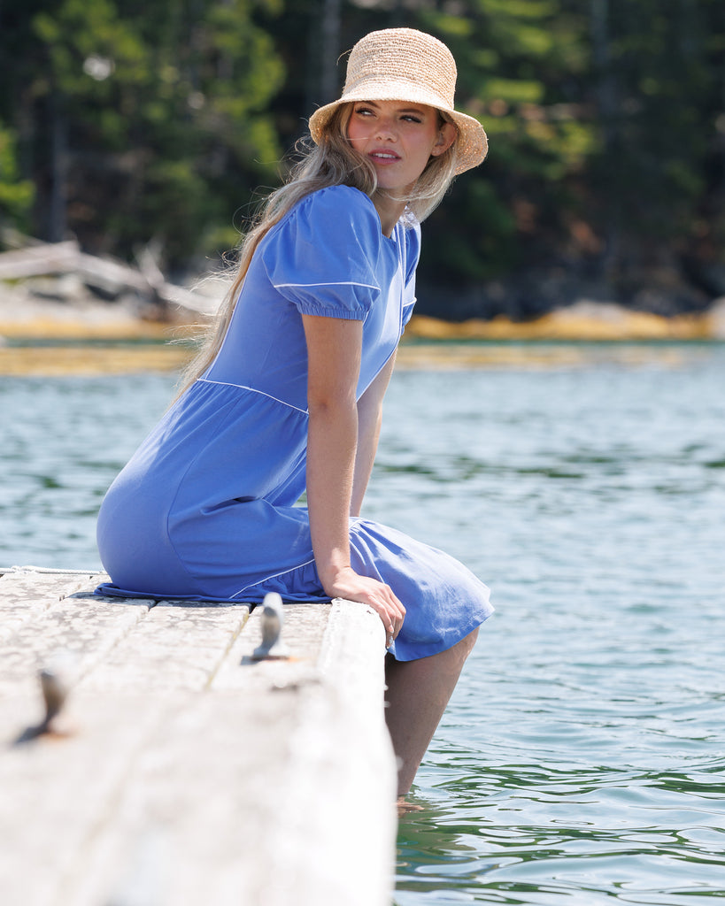 Woman in a blue dress and straw hat sitting by a body of water with trees in the background