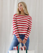 A woman wearing a red and off-white horizontally striped long-sleeve top with a wide boat neckline, paired with blue jeans, standing against a light neutral curtain background and holding a bag filled with gifts.