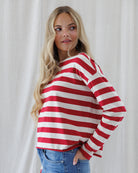 A woman wearing a red and off-white horizontally striped long-sleeve top with a wide boat neckline, paired with blue jeans, standing against a light neutral curtain background.