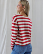 Back view of a woman wearing a red and off-white horizontally striped long-sleeve top with a wide boat neckline, paired with blue jeans, standing against a light neutral curtain background.
