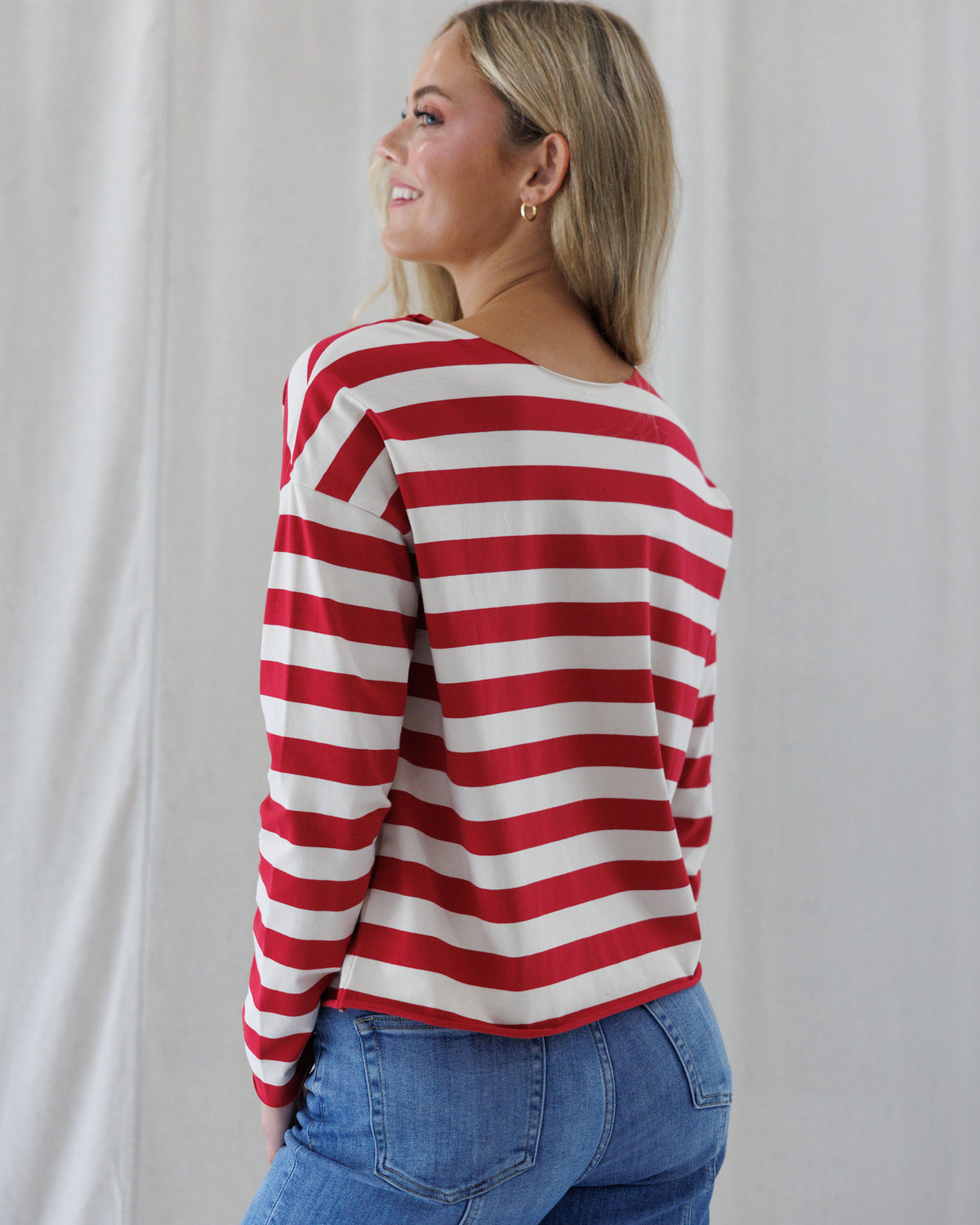 Back view of a woman wearing a red and off-white horizontally striped long-sleeve top with a wide boat neckline, paired with blue jeans, standing against a light neutral curtain background.