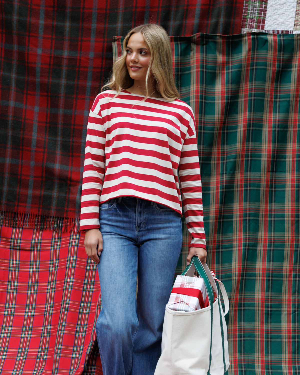 A woman wearing a red and white striped long-sleeve knit sweater and blue jeans stands in front of red and green plaid fabric backdrops, holding a white tote bag.