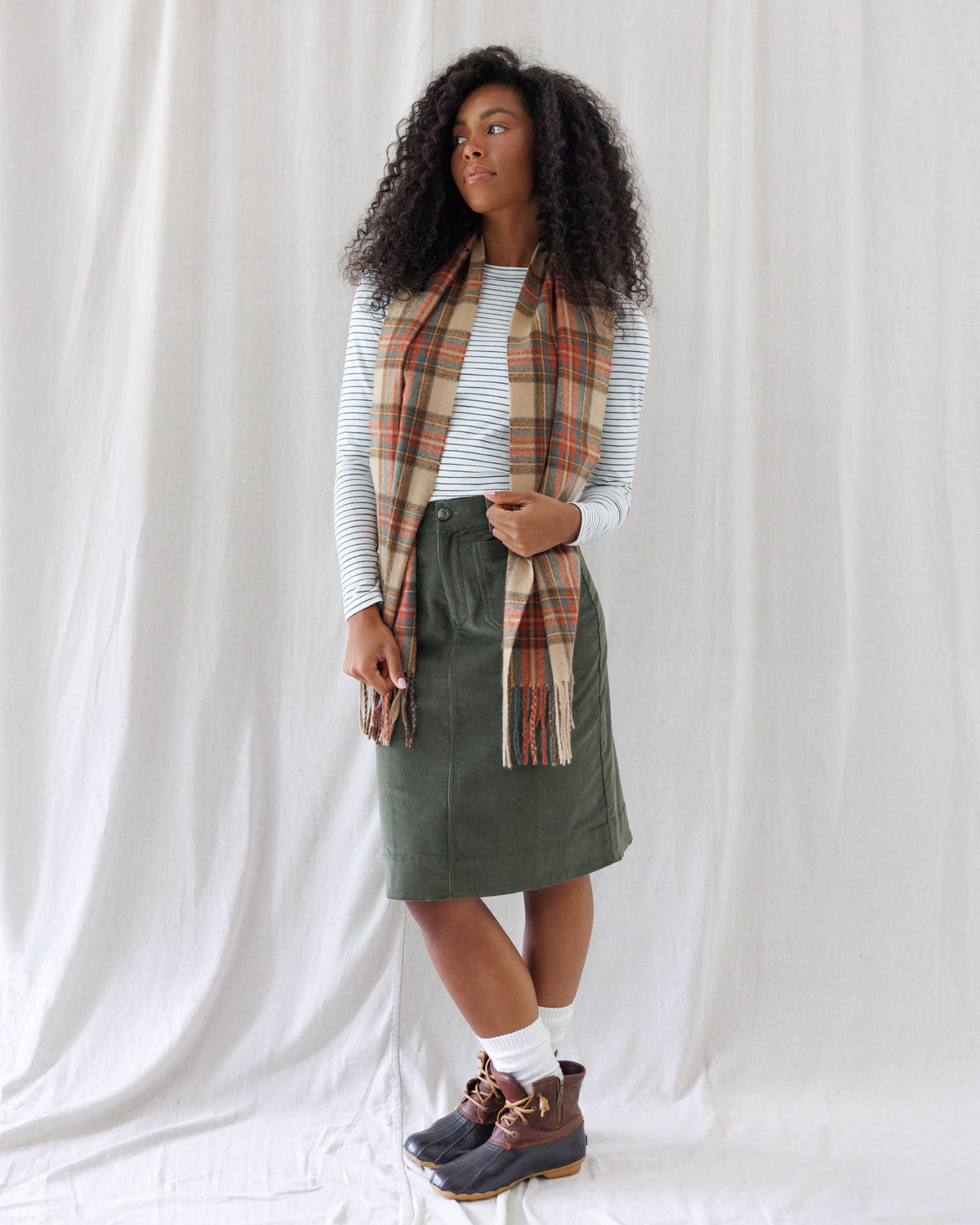 Model wearing a slim-fit white long-sleeve tee with narrow navy horizontal stripes and a classic crew neckline, styled tucked into an olive skirt with a plaid scarf.