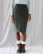 Front view of an olive green corduroy knee-length skirt featuring a high-rise waist, button fly, belt loops, and deep front patch pockets
