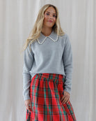 Front view of a heather grey knit crewneck sweater featuring a wide statement collar edged with delicate cream crochet trim, styled tucked into a red plaid skirt.
