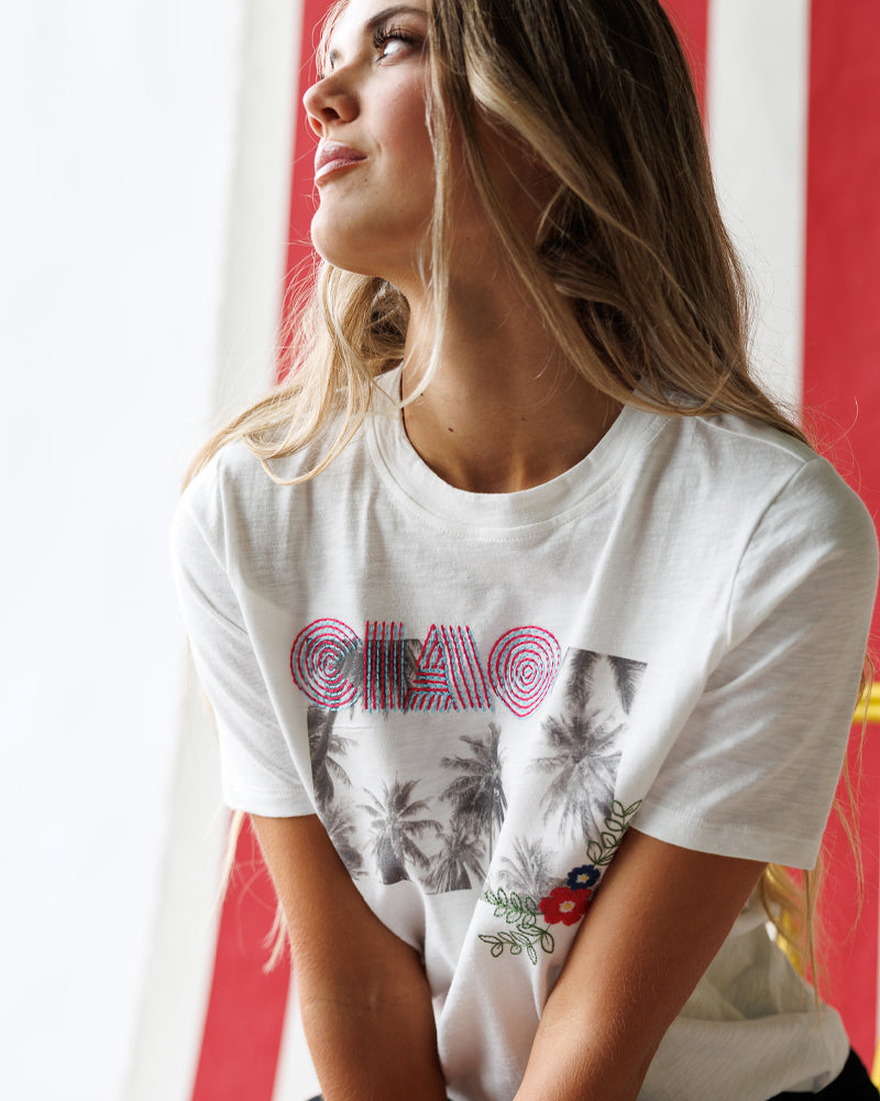 Model wearing a white short sleeve graphic tee with "ciao" and a summer scene 
